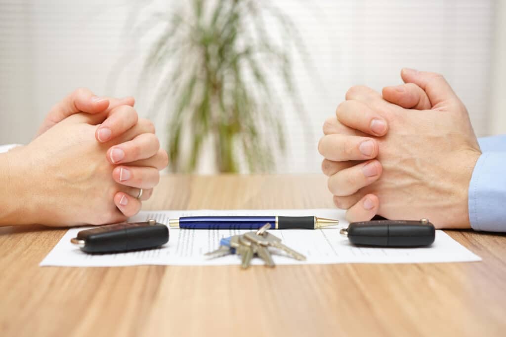 Two people seated across a table during divorce proceedings, with documents and house and car keys representing the division of matrimonial assets.